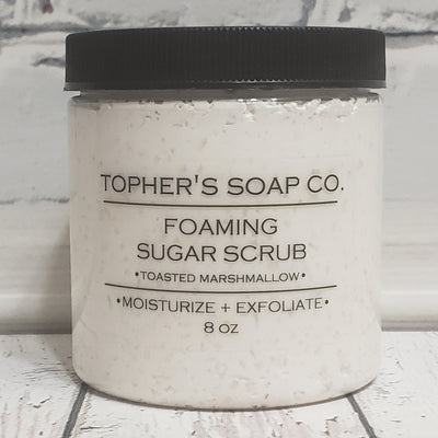 Off white sugar scrub in a clear jar with a black lid against a white brick background