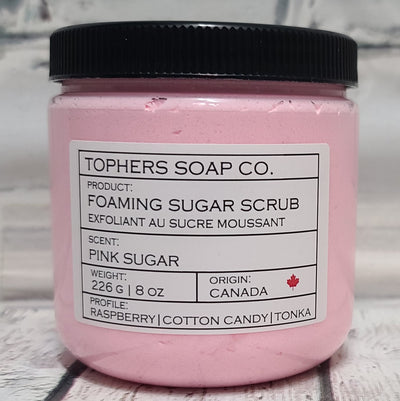 Light Pink  sugar scrub in a clear jar with a black lid against a white brick background