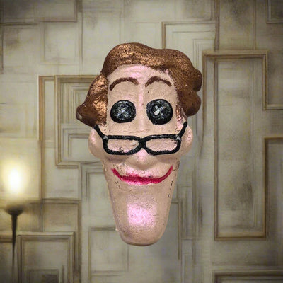 Topher’s Beard and Soap Co. Other Father Bath Bomb – hand-painted Coraline-inspired bath bomb displayed with a warped wood background and soft, muted wall sconce lighting to evoke the eerie, distorted charm of the Other World.