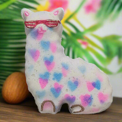 A hand-painted bath bomb shaped like a white llama wearing pink sunglasses, decorated with blue and pink hearts. It stands on a wooden surface with a blurred background of tropical leaves and flowers.