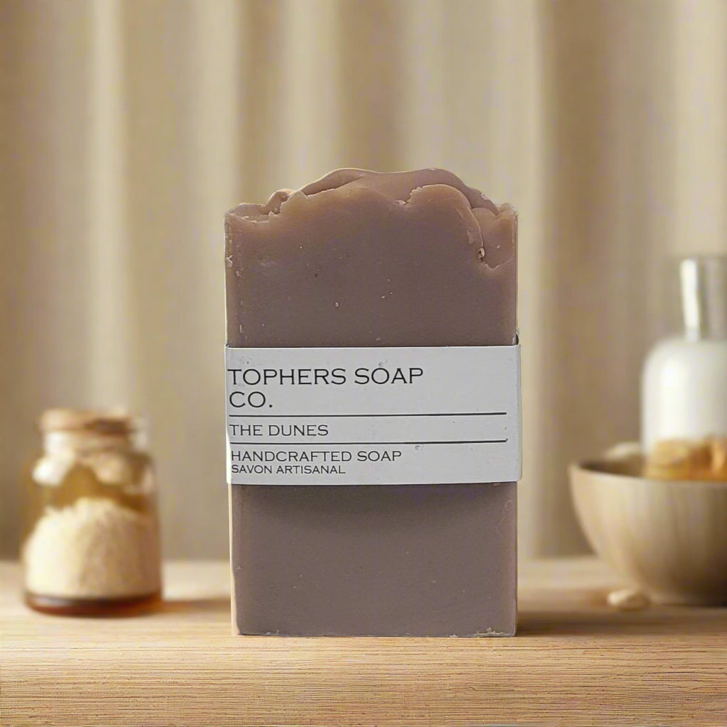 Topher's Beard and Soap Co. The Dunes Handcrafted Soap – muted mauve cold process soap bar with a minimalist label, styled on a wooden surface with natural spa elements like oats, honey, and ceramic vessels in a warm neutral setting