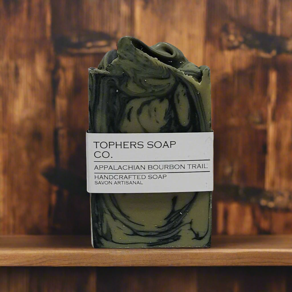 Topher's Beard and Soap Co. Appalachian Bourbon Trail Handcrafted Soap – rustic green and black swirled cold process soap bar with minimalist label, displayed on a wooden shelf with a charred woodgrain backdrop for a rugged, bourbon-inspired vibe