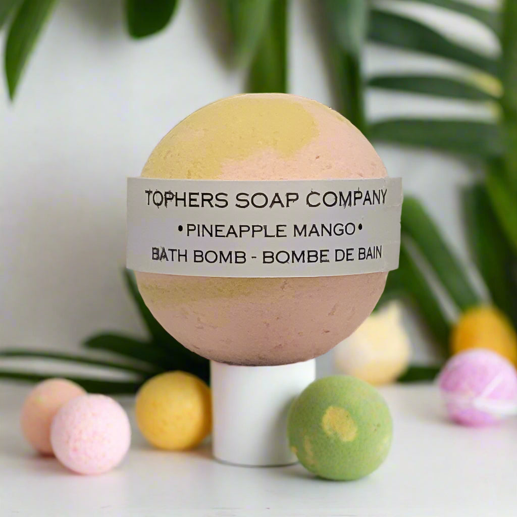 Topher's Beard and Soap Co. Pineapple Mango Bath Bomb – tropical yellow and peach spherical bomb with label, surrounded by mini bath bombs and lush greenery for a vibrant, fruity vibe