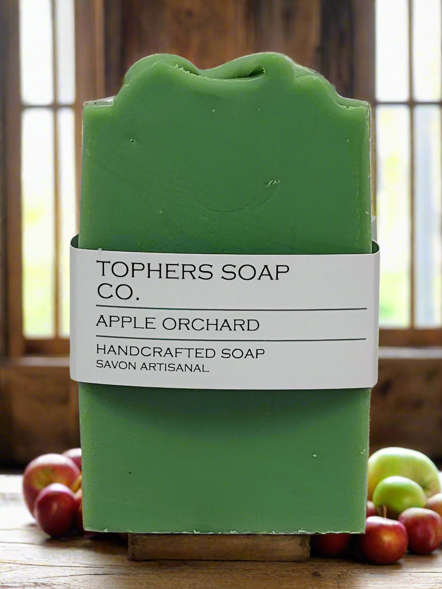 Topher's Beard and Soap Co. Apple Orchard Handcrafted Soap – vibrant green cold process soap bar with minimalist label, displayed on a rustic wood surface surrounded by fresh apples and a sunlit orchard window backdrop