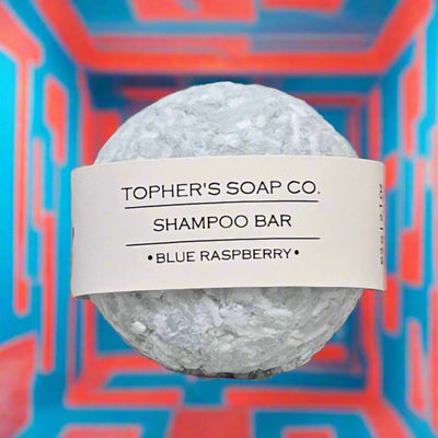 Topher's Beard and Soap Co. Blue Raspberry Shampoo Bar – round textured solid shampoo bar with minimalist label, displayed against a bold geometric red and blue background for a vibrant, modern aesthetic