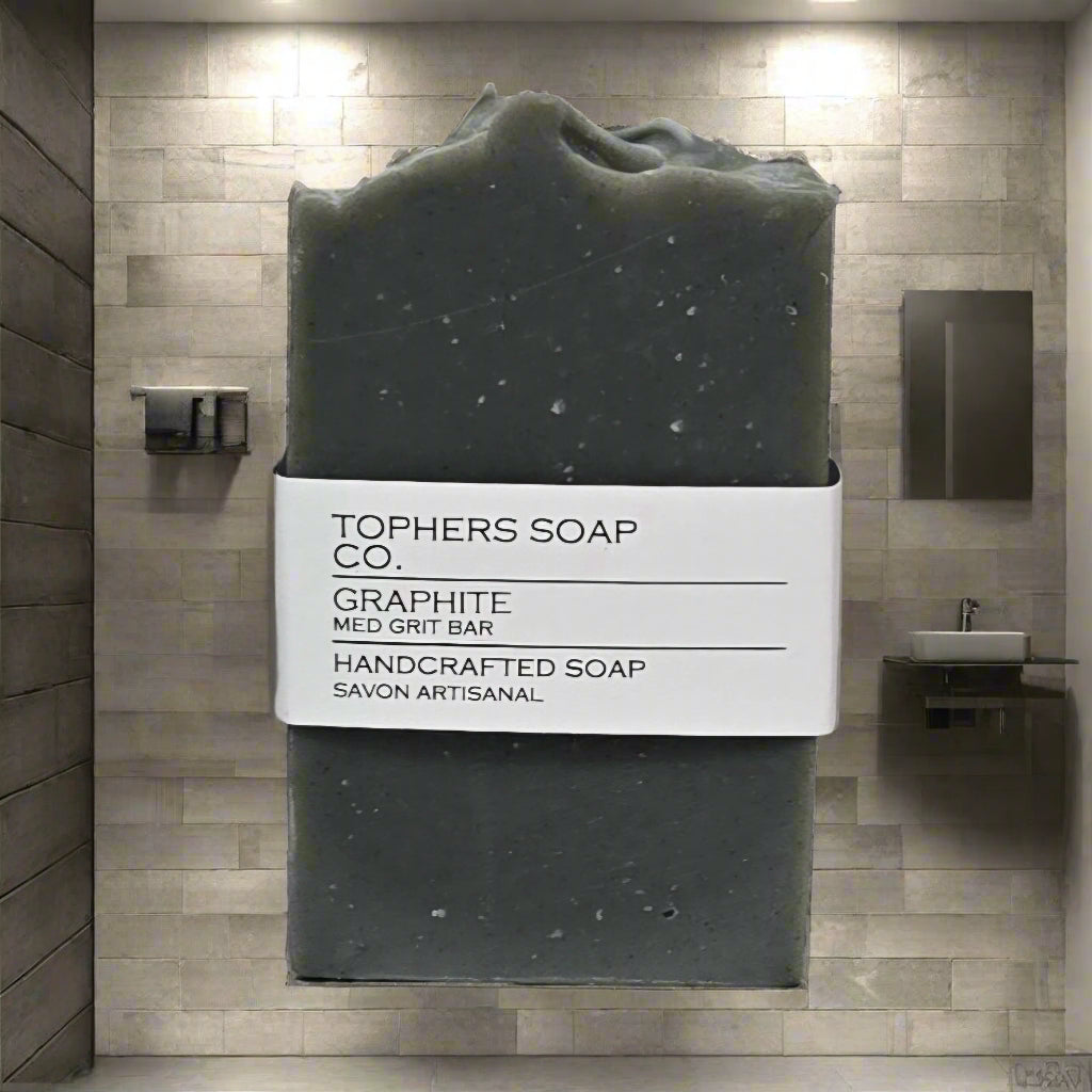 Topher's Beard and Soap Co. Graphite Handcrafted Soap – charcoal-grey exfoliating soap bar labeled as 'med grit', displayed in a sleek, modern tiled bathroom with moody lighting.