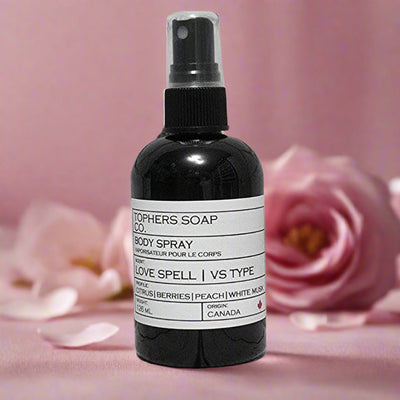"Topher's Beard and Soap Co. Love Spell Body Spray – 126 ml black bottle labeled with citrus, berries, peach, and musk scent profile, displayed on a soft pink background with rose petals and flowers