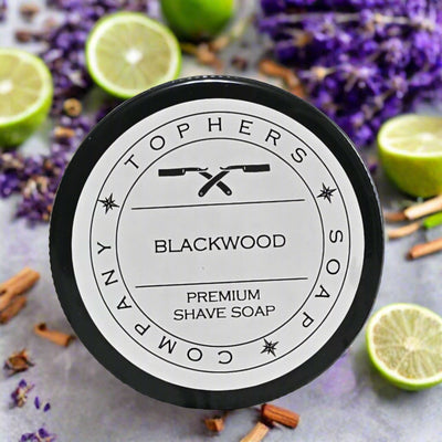 Topher's Beard and Soap Co. Blackwood Premium Shave Soap – top view of round tin with straight razor logo, surrounded by lavender, lime slices, and sandalwood pieces on a stone background