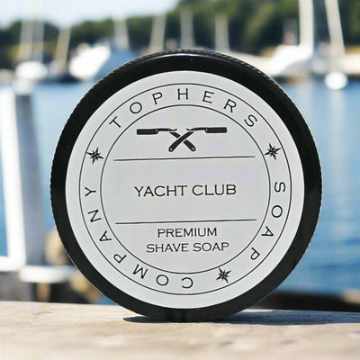 Yacht Club Premium Shave Soap by Topher's Beard and Soap Co. displayed on a dock with blurred sailboats and water in the background, evoking a luxurious seaside grooming experience.