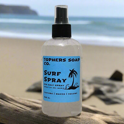 Topher’s Beard and Soap Co. Surf Spray Sea Salt Spray – 268 ml beachy texture spray with a blue label, displayed on driftwood at the beach with ocean waves and sand in the background, styled to evoke volume, waves, and summer hair vibes.
