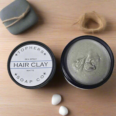 Topher’s Beard and Soap Co. Sea Spray Hair Clay – matte-finish styling clay shown in an open tin beside its lid, styled on a clean wood surface with beach-inspired accents like a twine loop, smooth white stones, and a wrapped gift box for a natural, coastal grooming vibe.

