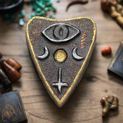 Topher’s Beard and Soap Co. Ouija Board Planchette Bath Bomb – hand-painted bath bomb shaped like a Ouija planchette, displayed on a scarred wooden tabletop surrounded by voodoo and magik items, styled to evoke a mystical, ritualistic atmosphere.