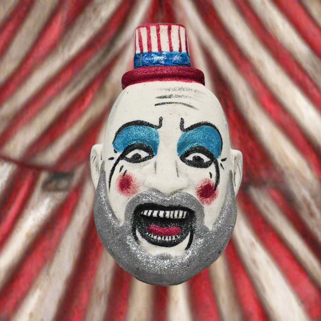 Topher’s Beard and Soap Co. Captain Spaulding Bath Bomb – hand-painted horror-inspired bath bomb depicting the character from Rob Zombie’s films, displayed against a filthy red and white circus tent backdrop to evoke a gritty, unsettling carnival atmosphere.