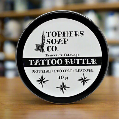 Round black tin of Tophers Soap Co. Tattoo Butter, featuring a black and white label with "TATTOO BUTTER" prominently displayed, along with "Nourish | Protect | Restore" and "30 g," photographed on a wooden surface, emphasizing a locally made product from Brantford, Ontario, designed to care for tattoos.