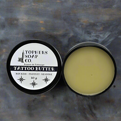 Two round silver tins of Tophers Soap Co. Tattoo Butter, one closed showing the black and white label with "TATTOO BUTTER" and "Nourish | Protect | Restore," and the other open revealing the light yellow balm inside, emphasizing a locally handcrafted product from Brantford, Ontario, designed to nourish and protect tattoos.