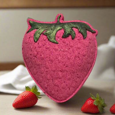 A strawberry-shaped bath bomb, Hand painted by Tophers Soap Co. textured and deep pink with dark green, shimmery leaf details at the top, is the central focus. It hovers slightly above a light gray surface. Three fresh strawberries with green stems and leaves are scattered around the base of the bath bomb. The background is softly blurred, showing a light brown wooden surface and a hint of white fabric or a container. The lighting is soft and highlights the color of the bath bomb and the real strawberries.