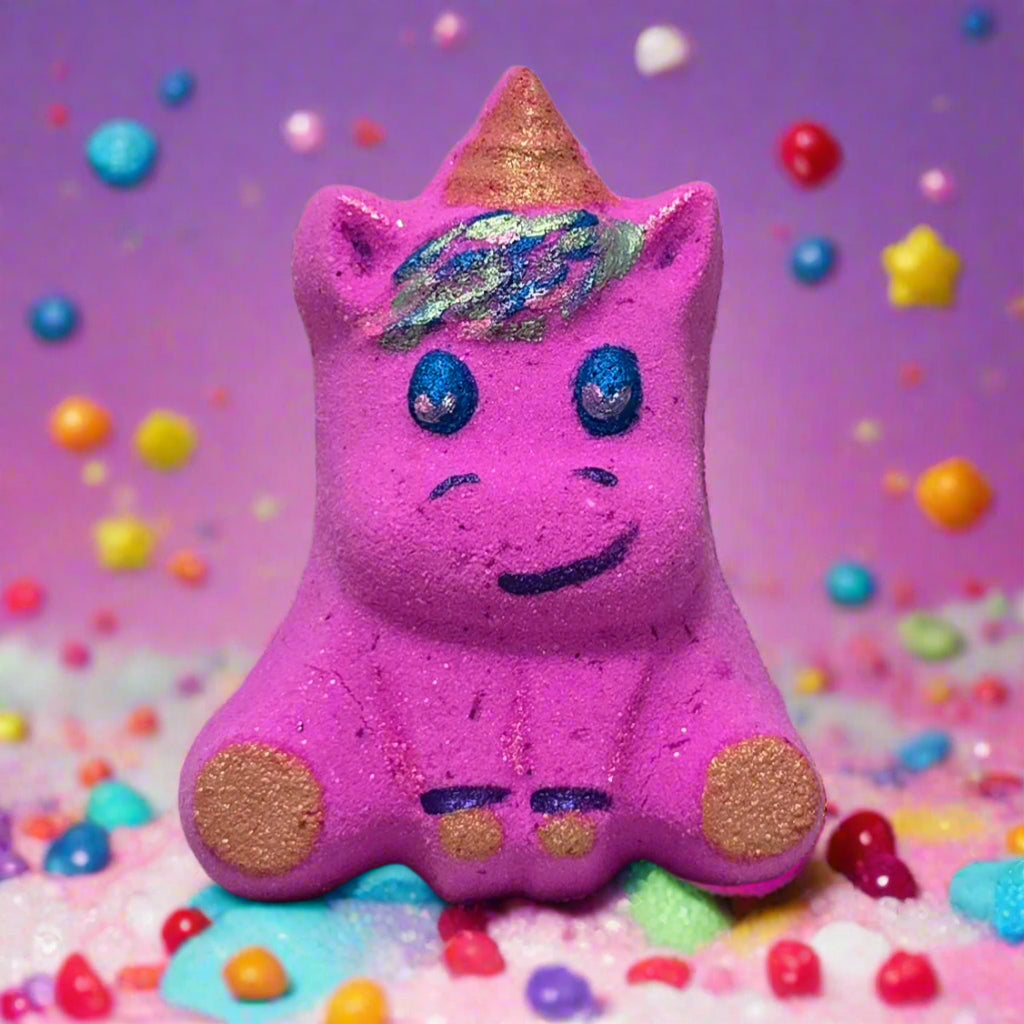 A purple, glittery unicorn-shaped bath bomb with a golden horn, blue eyes, and a smiling face sits on a bed of colorful bath crystals against a purple background with floating pastel spheres. Tophers Soap Co.