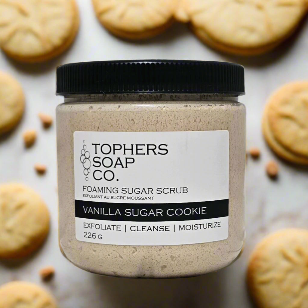 Topher's Soap Co. Foaming Sugar Scrub in a clear jar with a black lid. The scrub is a light beige color and the label clearly reads "VANILLA SUGAR COOKIE" and "EXFOLIATE | CLEANSE | MOISTURIZE" along with the weight "226 G." The jar is surrounded by vanilla sugar cookies, emphasizing the sweet and comforting fragrance. Handcrafted in Brantford, Ontario, Canada.