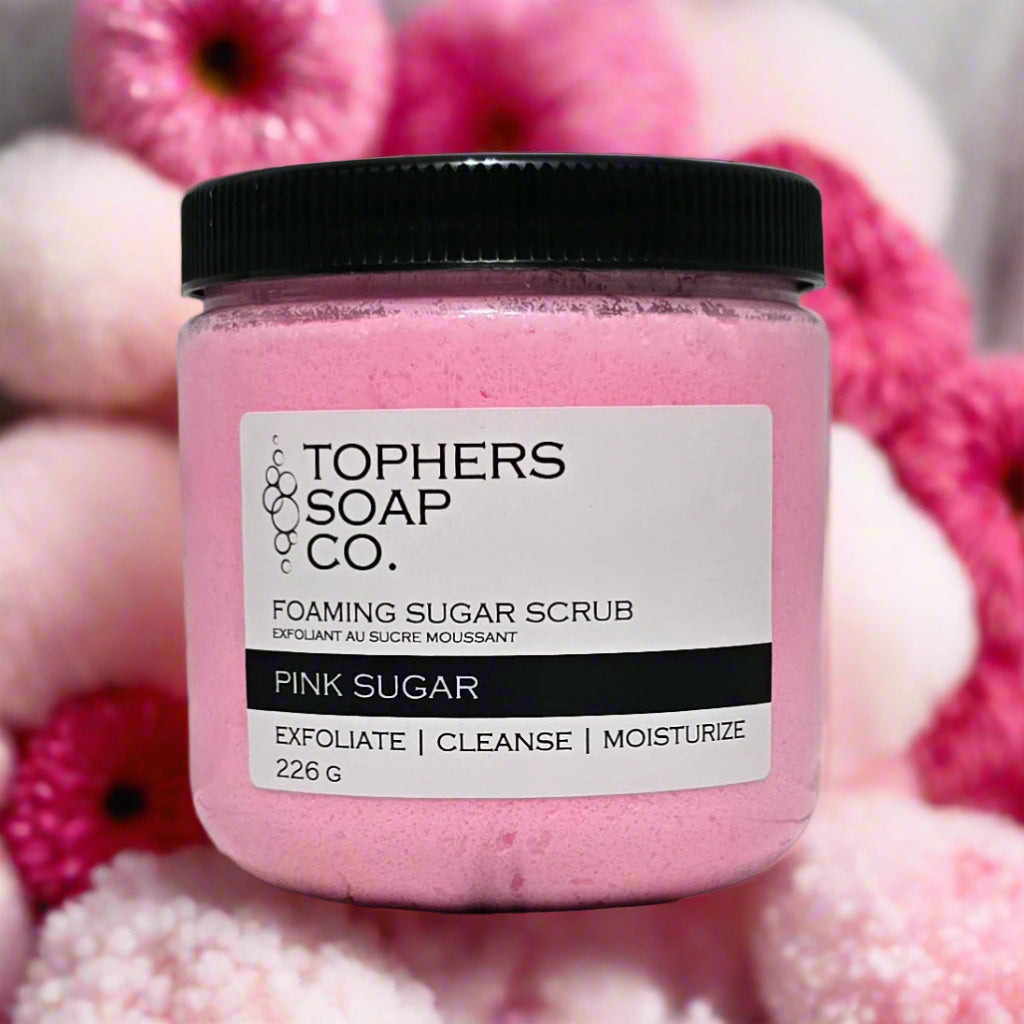 Topher's Soap Co. Foaming Sugar Scrub in a clear jar with a black lid. The scrub is a vibrant pink color and the label clearly reads "PINK SUGAR" and "EXFOLIATE | CLEANSE | MOISTURIZE" along with the weight "226 G." The jar is surrounded by blurred pink and white flowers, emphasizing the sweet and delightful fragrance. Handcrafted in Brantford, Ontario, Canada.