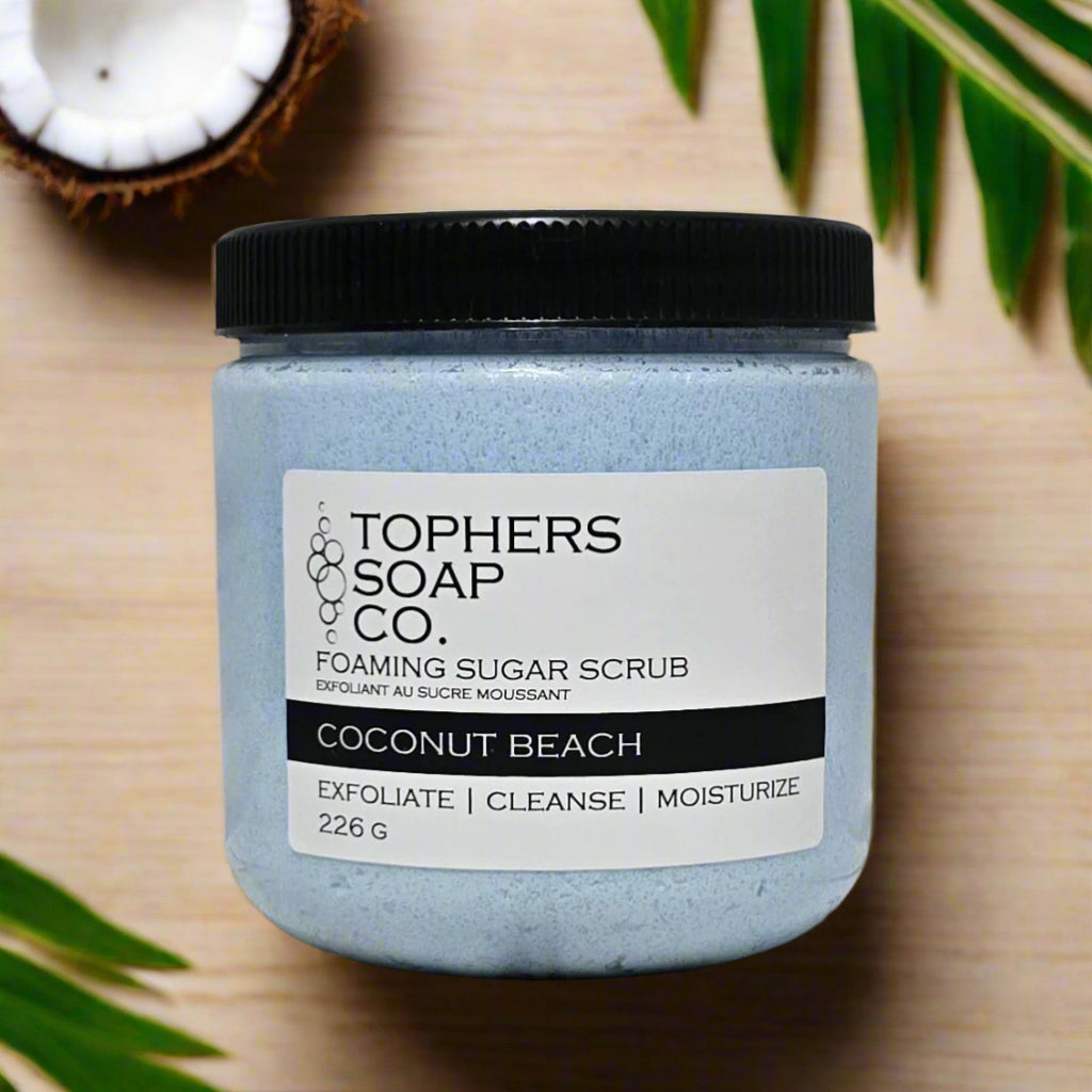 Topher's Soap Co. Foaming Sugar Scrub in a clear jar with a black lid. The scrub is a light blue color and the label clearly reads "COCONUT BEACH" and "EXFOLIATE | CLEANSE | MOISTURIZE" along with the weight "226 G." The jar is surrounded by a half of a coconut and green tropical leaves on a light wooden surface, emphasizing the tropical coconut scent. Handcrafted in Brantford, Ontario, Canada.
