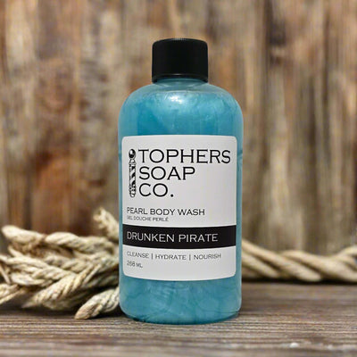 A clear plastic bottle with a black cap containing a shimmering light blue liquid, identified by a white label as "TOPHER'S SOAP CO. PEARL BODY WASH GEL DOUCHE PERLÉ DRUNKEN PIRATE." The label also indicates "CLEANSE | HYDRATE | NOURISH" and "268 mL." The bottle is positioned in front of a blurred background featuring a thick rope.