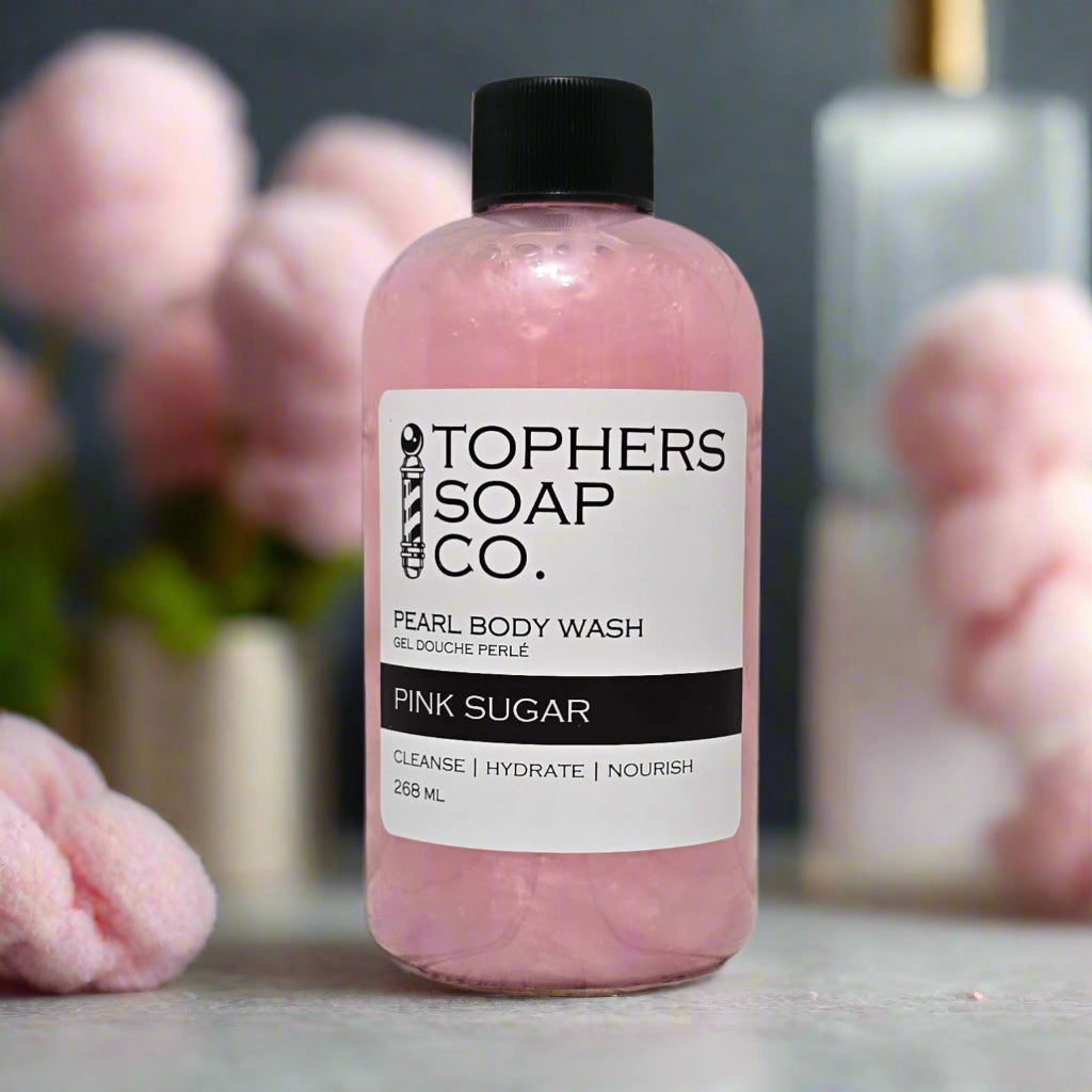 A clear plastic bottle with a black cap containing a shimmering light pink liquid, identified by a white label as "TOPHER'S SOAP CO. PEARL BODY WASH GEL DOUCHE PERLÉ PINK SUGAR." The label also indicates "CLEANSE | HYDRATE | NOURISH" and "268 mL." The bottle is positioned on a light gray surface with blurred pink, fluffy elements in the background.
