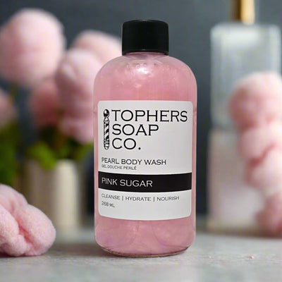 A clear plastic bottle with a black cap containing a shimmering light pink liquid, identified by a white label as "TOPHER'S SOAP CO. PEARL BODY WASH GEL DOUCHE PERLÉ PINK SUGAR." The label also indicates "CLEANSE | HYDRATE | NOURISH" and "268 mL." The bottle is positioned on a light gray surface with blurred pink, fluffy elements in the background.