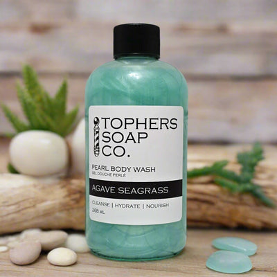 A clear plastic bottle with a black cap containing a shimmering light teal liquid, identified by a white label as "TOPHER'S SOAP CO. PEARL BODY WASH GEL DOUCHE PERLÉ AGAVE SEAGRASS." The label also indicates "CLEANSE | HYDRATE | NOURISH" and "268 mL." The bottle is positioned on a light-colored surface with small rocks and a piece of driftwood. A blurred background shows a wooden surface and small potted plants.