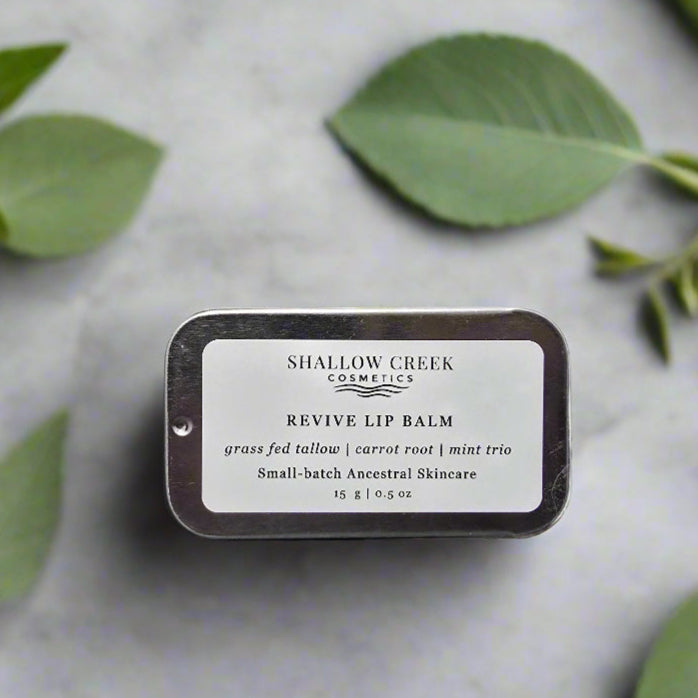 Shallow Creek Cosmetics Revive Lip Balm, a 15g tin of grass-fed tallow, carrot root, and mint trio balm, presented on a light background with green leaves.