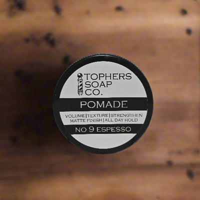 
A top-down view of a black jar of Topher's Soap Co. No. 9 Espresso pomade. The white circular label prominently features the brand's barber pole logo, "TOPHERS SOAP CO." and "POMADE." Below this, "VOLUME | TEXTURE | STRENGTHEN" and "MATTE FINISH | ALL DAY HOLD" are listed in smaller text, with "NO 9 ESPESSO" at the bottom of the label. The background is a blurred wooden surface.
