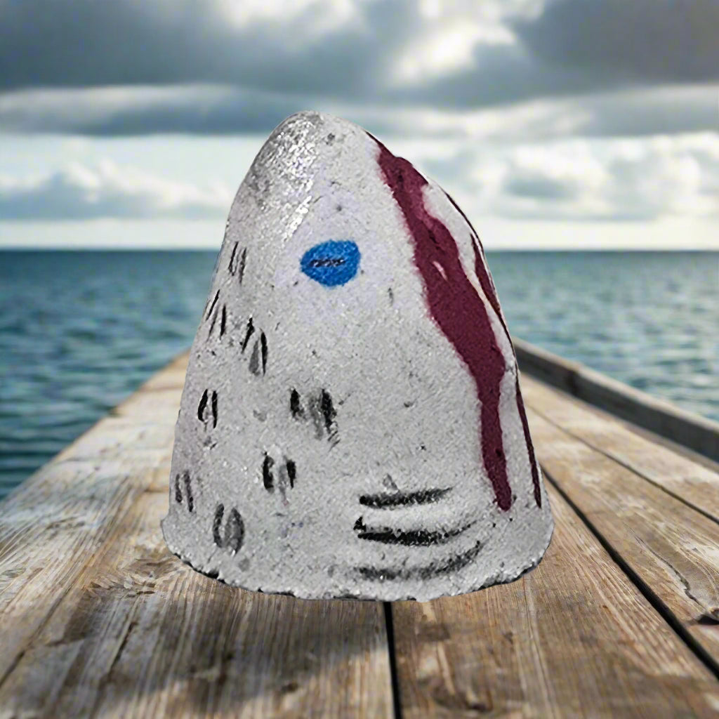 Shark Bite Bath Bomb by Topher's Beard and Soap Co., hand-painted with blood splatter detail, displayed on a weathered dock with the ocean and cloudy sky in the background.