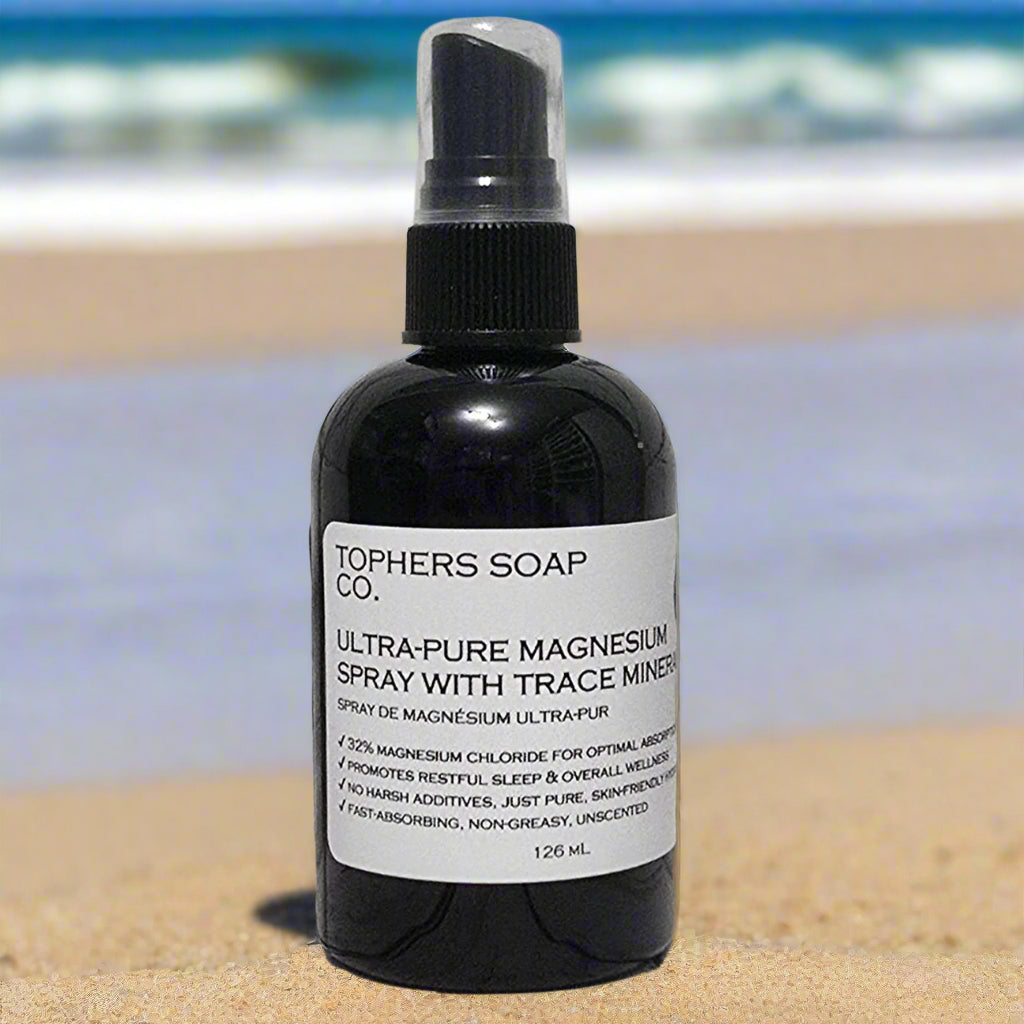 Tophers Soap Co. Ultra-Pure Magnesium Spray bottle standing upright on warm beach sand with gentle ocean waves and soft sky in the background.