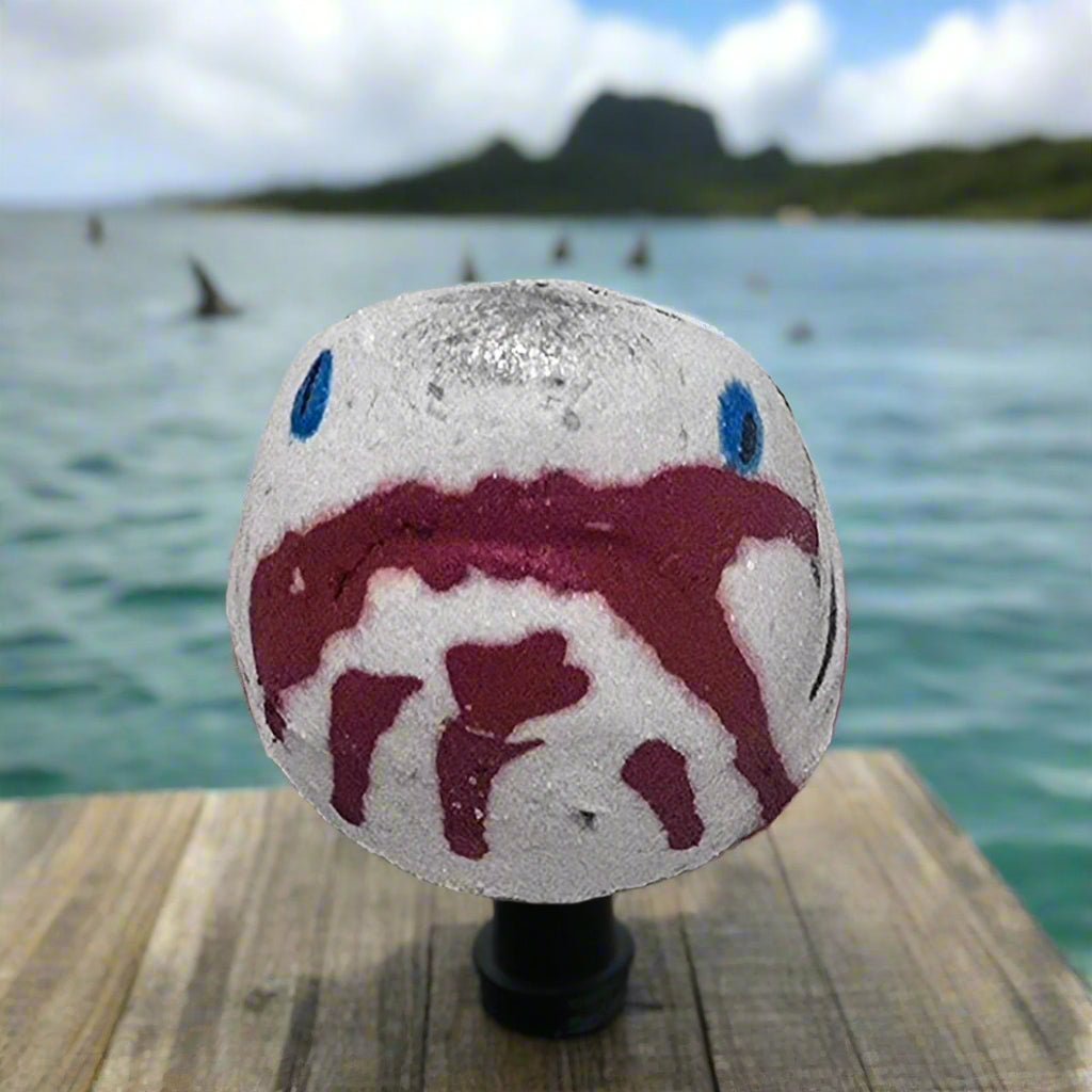 Shark Bite Bath Bomb by Topher's Beard and Soap Co., hand-painted with blood-red detail, displayed on a wooden dock with ocean, mountains, and dorsal fins in the distance.