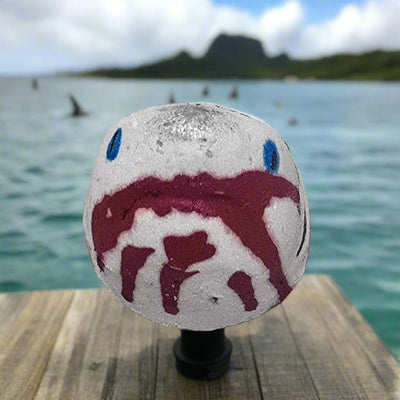 Shark Bite Bath Bomb by Topher's Beard and Soap Co., hand-painted with blood-red detail, displayed on a wooden dock with ocean, mountains, and dorsal fins in the distance.