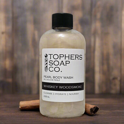 A clear plastic bottle with a black cap containing a light beige, shimmering liquid, identified by a white label as "TOPHER'S SOAP CO. PEARL BODY WASH GEL DOUCHE PERLÉ WHISKEY WOODSMOKE." The label also indicates "CLEANSE | HYDRATE | NOURISH" and "268 mL." The bottle is positioned on a wooden surface with two cinnamon sticks placed near it. A blurred wooden background is visible.