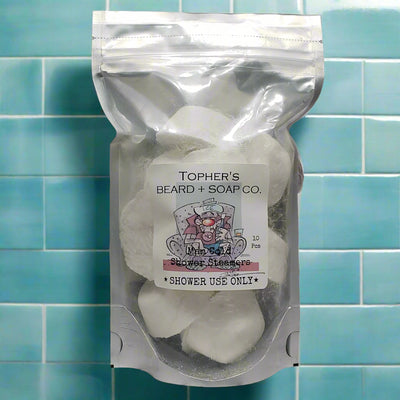 A clear, resealable silver pouch contains ten white shower steamers labeled "TOPHER'S BEARD + SOAP CO." The label features a cartoon bull relaxing in a chair and reads "Man Cold Shower Steamers" and "SHOWER USE ONLY". The pouch is placed against a background of light blue and white square tiles.