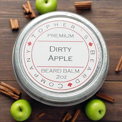 A top-down view of a round, silver tin of beard balm. The white label on the lid reads "TOPHER'S BEARD COMPANY" in a circular arrangement around the edge, interspersed with small red maple leaves. In the center, "PREMIUM" is at the top, followed by "Dirty Apple" in a larger font, and "BEARD BALM 2oz" below. The tin is surrounded by three green apples and several cinnamon sticks arranged on a dark brown wooden surface.