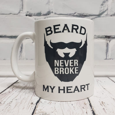 White 11 oz ceramic mug from Tophers Beard Co. in Brantford, Ontario, featuring a black graphic of a beard and the text "BEARD NEVER BROKE MY HEART" printed using a durable sublimation method.

