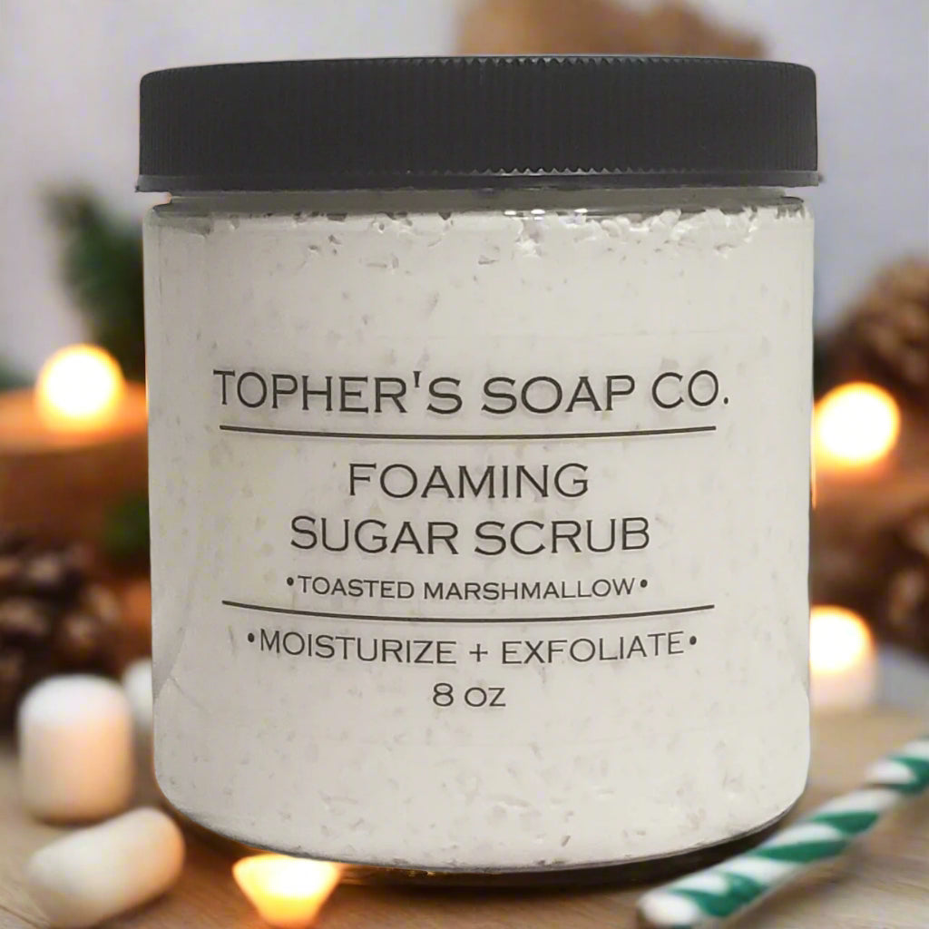 Jar of Tophers Soap Co. Toasted Marshmallow foaming sugar scrub, a light, textured scrub with a black lid, surrounded by marshmallows, pinecones, and soft lights, evoking a cozy atmosphere and highlighting its Brantford, Ontario origin.