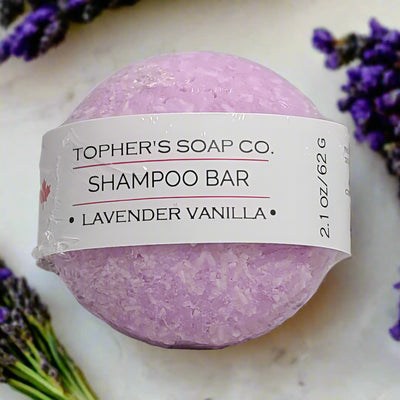 A round, light purple Lavender Vanilla Shampoo Bar from Tophers Soap Co., wrapped in a white label with the product name and weight (2.1 oz / 62 g), surrounded by sprigs of fresh lavender.