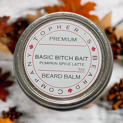A top-down view of a round, silver tin of beard balm. The white label on the lid reads "TOPHER'S BEARD COMPANY" in a circular arrangement around the edge. In the center, "PREMIUM" is at the top, followed by "BASIC BITCH BAIT" in a larger font, then "PUMPKIN SPICE LATTE" in smaller text, and "BEARD BALM" below. The weight "2oz" is also visible. A small Canadian maple leaf is incorporated into the design. The tin is surrounded by blurred autumn leaves and pinecones on a light wooden surface.