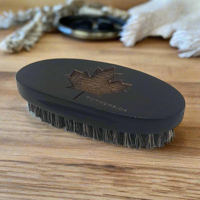 Angled view of the black, oval Boar Hair Brush from Tophers Beard Co., clearly showing the laser-cut Canadian maple leaf and "TOPHERS.CA" on the handle, emphasizing its availability in Brantford, ON