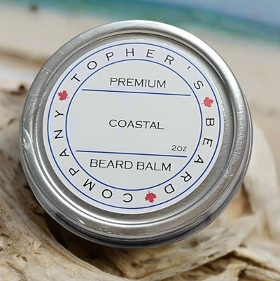 A top-down view of a silver tin containing Tophers Premium Coastal Beard Balm. The white label features the product name, "2oz," and small Canadian maple leaves. The tin rests on a piece of driftwood, with a blurred beach scene in the background.