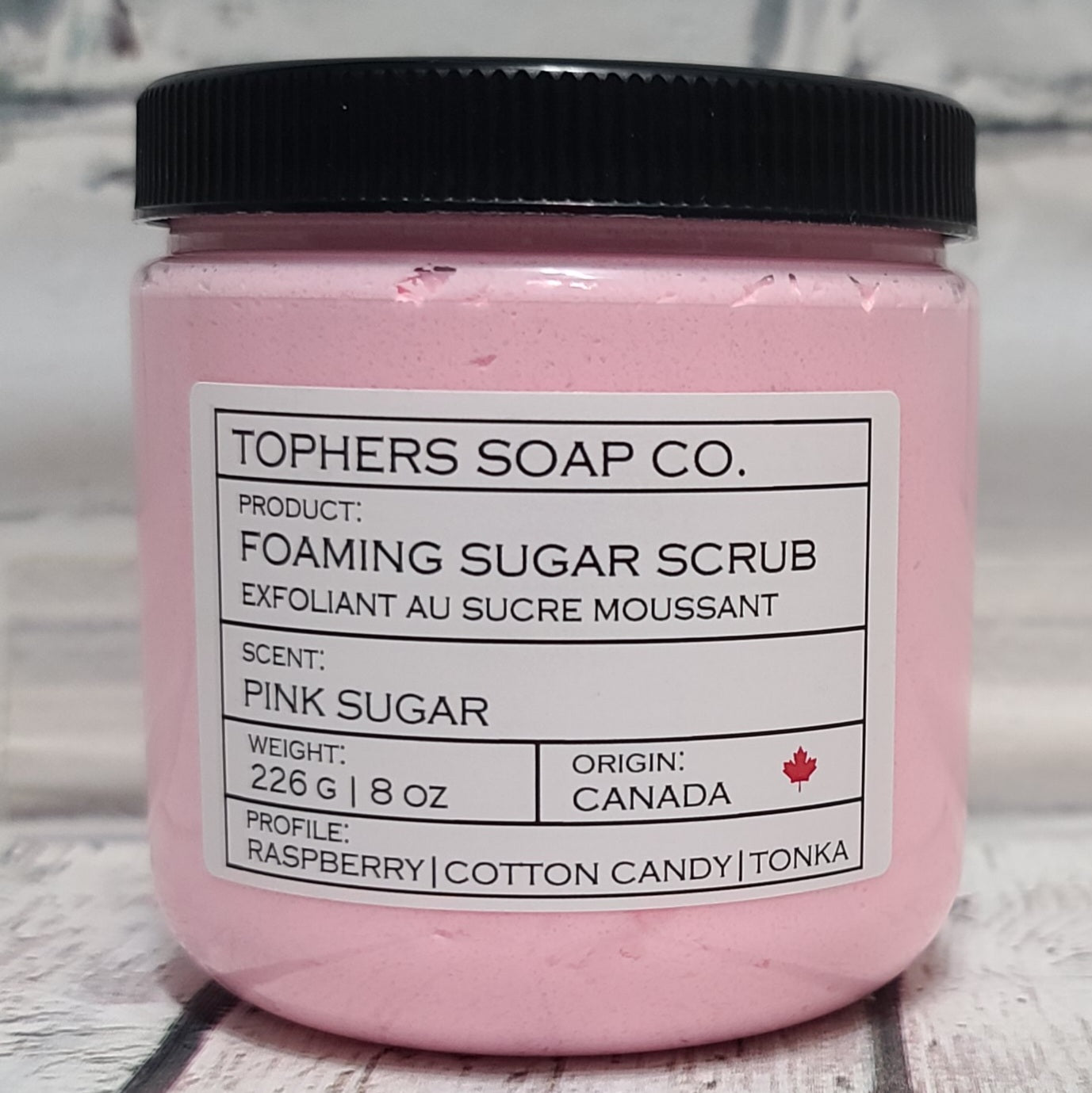 Light Pink  sugar scrub in a clear jar with a black lid against a white brick background