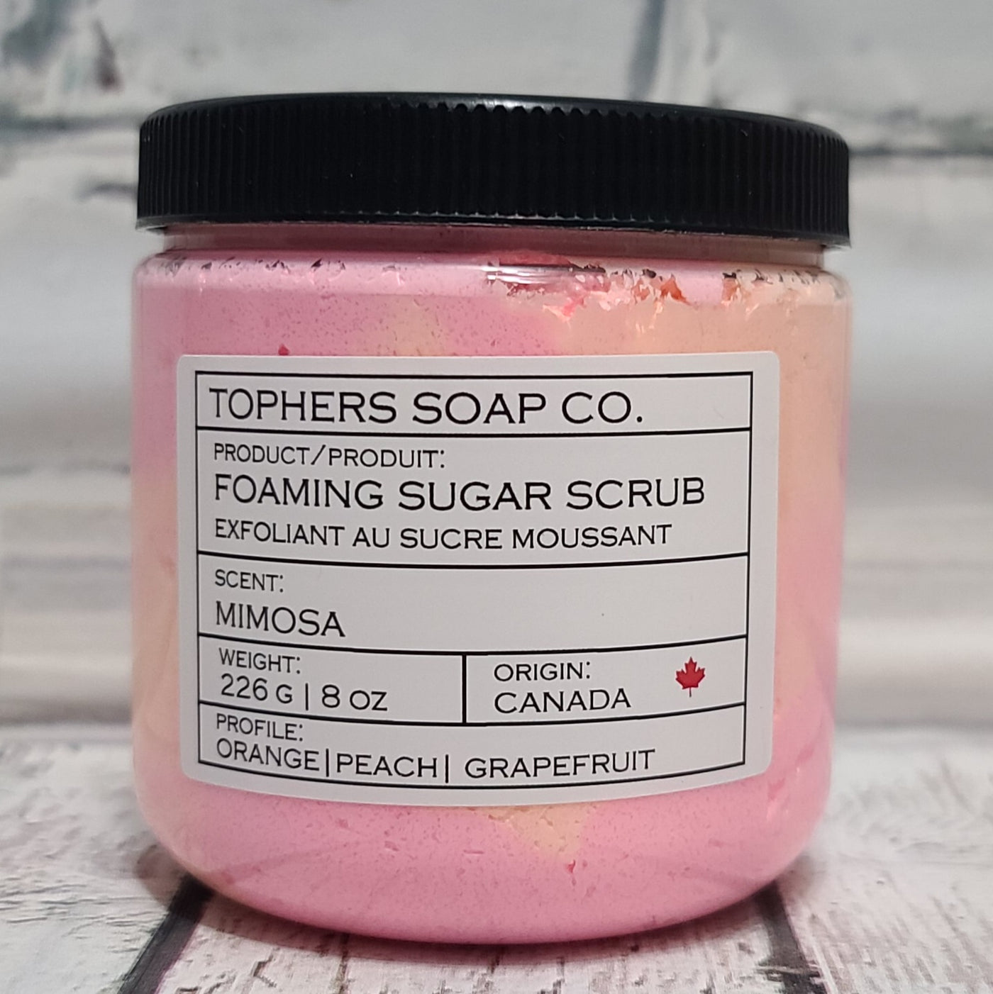 Orange and pink sugar scrub in a clear jar with a black lid against a white brick background