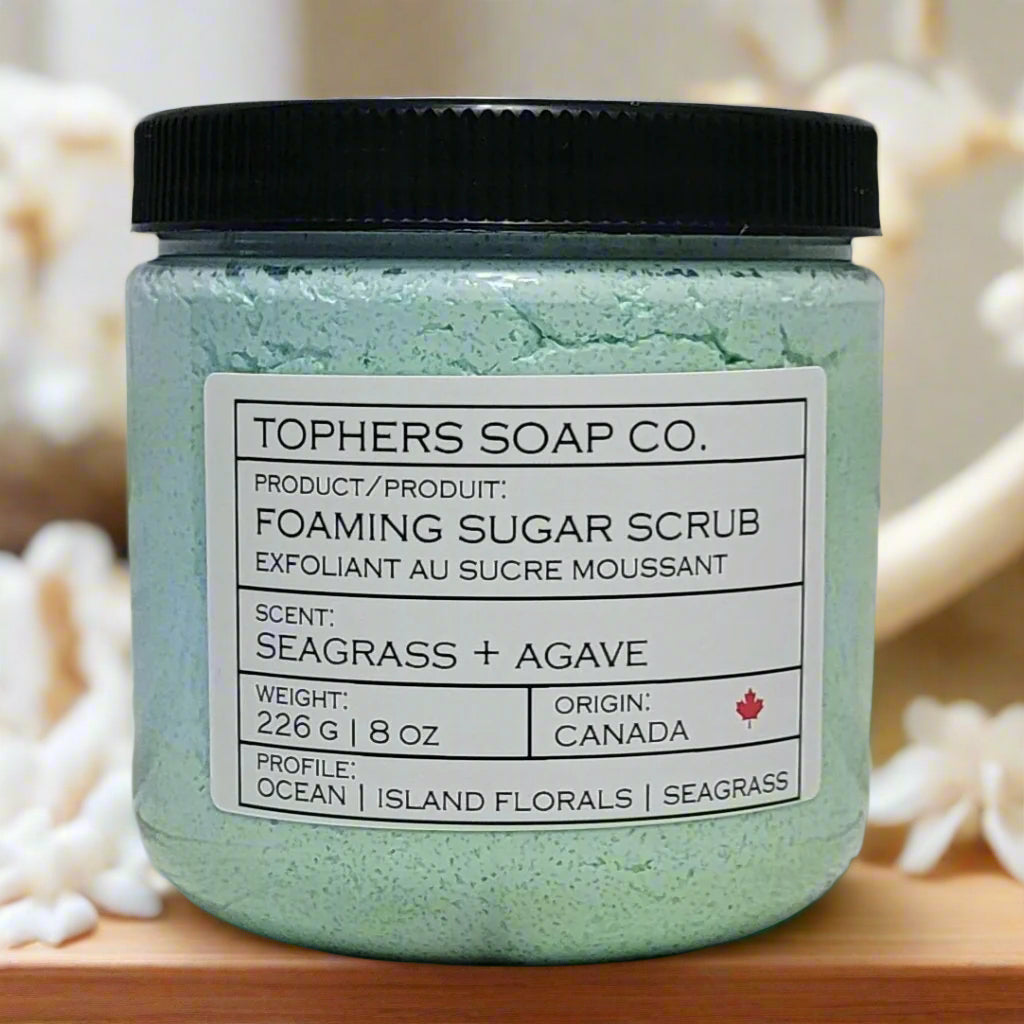 A clear plastic jar with a black lid contains Topher's Soap Co. Foaming Sugar Scrub in the scent "Seagrass + Agave." The label indicates a scent profile of ocean, island florals, and seagrass. The jar contains 226 g or 8 oz of product and states it originated in Canada. The background is blurred but shows light-colored, possibly sandy or shell-like textures.