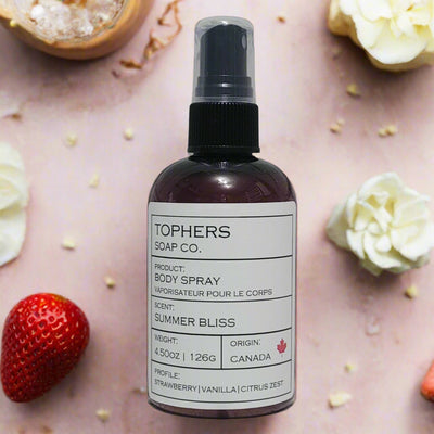 Dark bottle of Tophers Soap Co. Summer Bliss Body Spray with a clear spray nozzle and a white label detailing strawberry, vanilla, and citrus zest scent notes, surrounded by fresh strawberries and white flowers on a light pink background, emphasizing its Canadian origin in Brantford, Ontario.