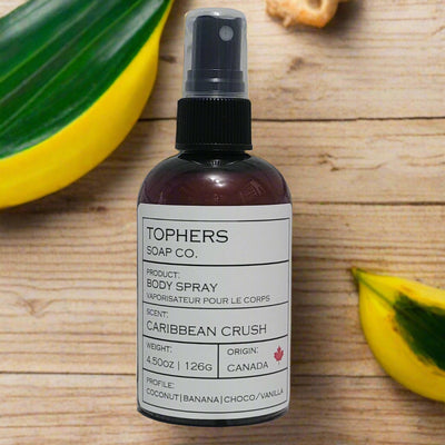 Dark bottle of Tophers Soap Co. Caribbean Crush Body Spray with a black spray nozzle and a white label detailing coconut, banana, chocolate, and vanilla scent notes, against a backdrop of tropical leaves on a wooden surface, emphasizing its Canadian origin in Brantford, Ontario.