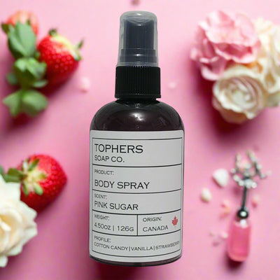 Dark bottle of Tophers Soap Co. Pink Sugar Body Spray with a clear spray nozzle and a white label detailing cotton candy, vanilla, and strawberry scent notes, surrounded by fresh strawberries and pink flowers on a pink background, emphasizing its Canadian origin in Brantford, Ontario.