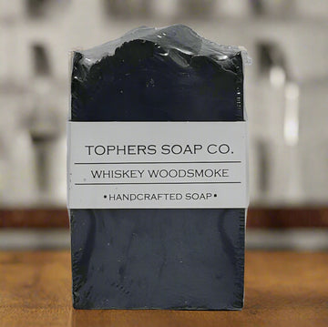 Bar of Tophers Soap Co. Whiskey Woodsmoke Handcrafted Soap, featuring a dark grey/black color, wrapped in a white label with the product name and "HANDCRAFTED SOAP," photographed on a wooden surface with blurred background elements, emphasizing a locally made (Brantford, Ontario, Canada) soap with a bold, smoky, and masculine scent, likely containing detoxifying activated charcoal.